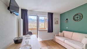 Ett rum med ett träbord, en soffa, en tv som är monterad på väggen och en utsikt över bergen genom fönstret.