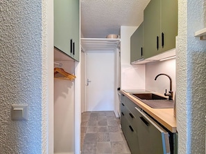 Een smalle keuken met groene kastjes, een witte deur en een betegelde vloer.