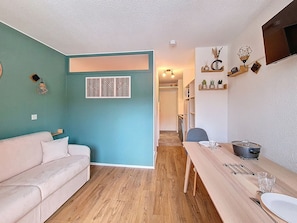 A compact living space with a teal accent wall, a wooden floor, a sofa, a dining table with chairs, and a kitchen area with a TV and decorative items. - Bright studio with south-facing balcony at the foot of the slopes (Val Thorens)
