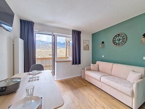 Ein Zimmer mit Esstisch, einer Couch, Blick auf Berge durch ein Fenster und einer Wanduhr.
