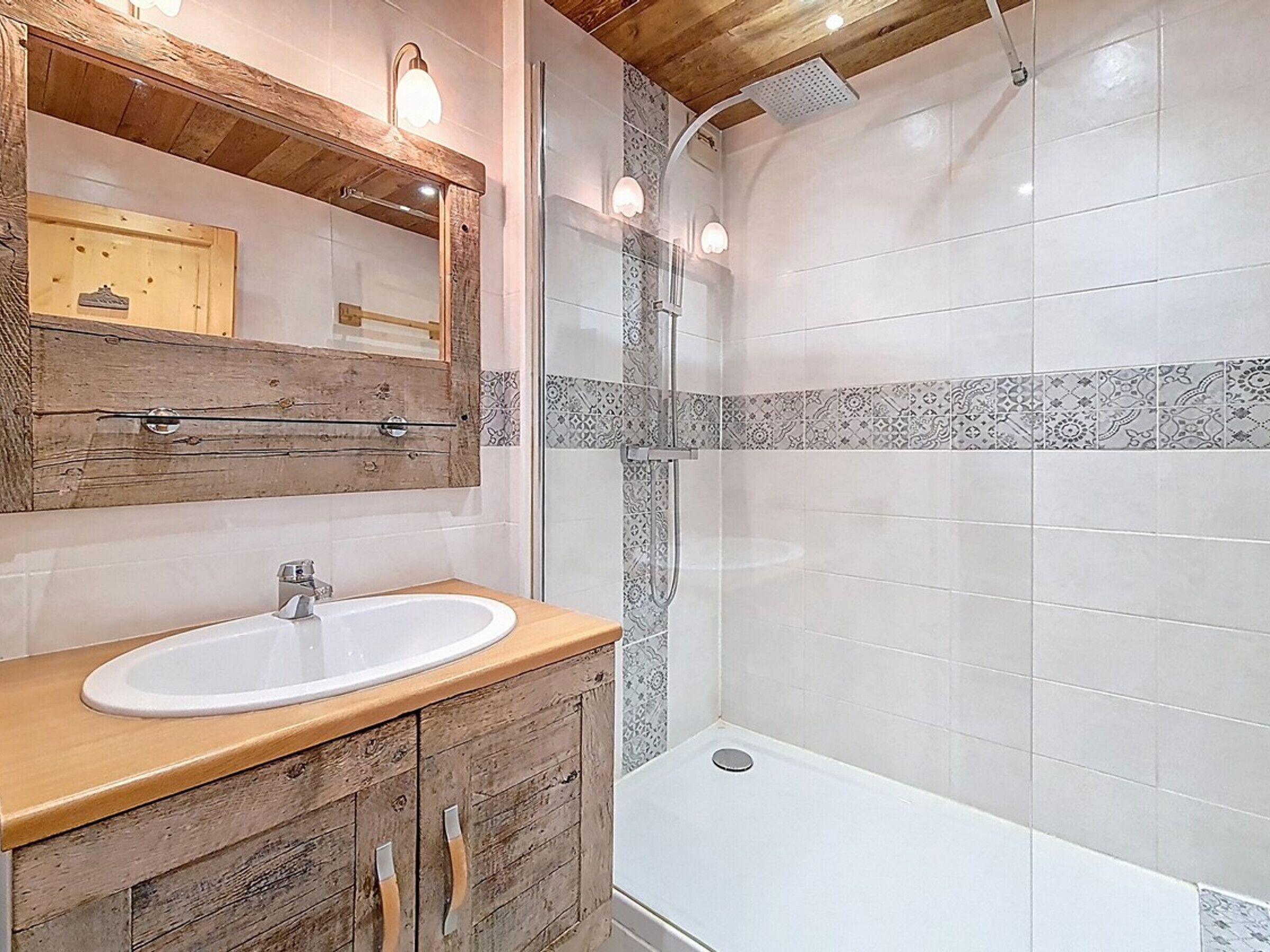 Un baño con tocador de madera, un lavabo blanco, una ducha con puerta de cristal y azulejos estampados.