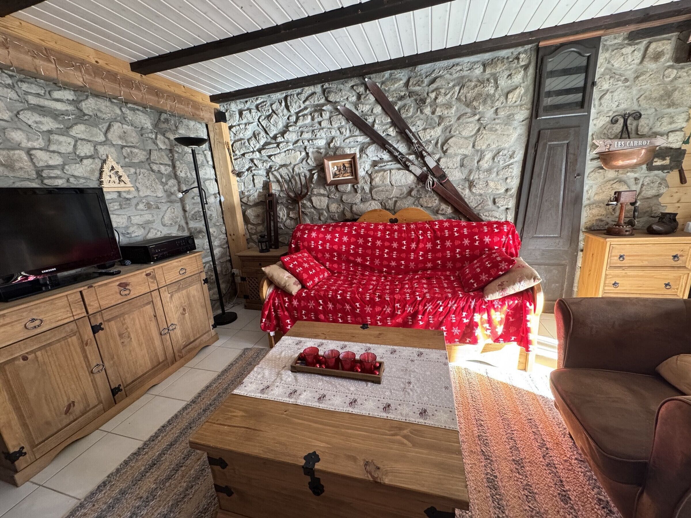 A cozy living room with a stone wall, wooden furniture, a red patterned sofa, and skis leaning against the wall.