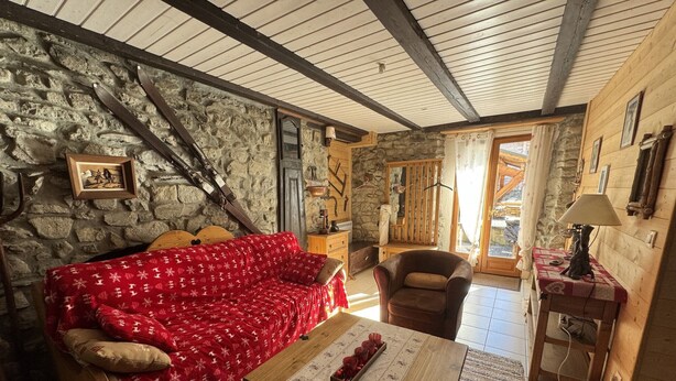 Une pièce chaleureuse avec un mur en pierre, un canapé rouge à motifs, une table en bois sur laquelle se trouve un plateau avec des tasses rouges, un fauteuil en cuir marron et un meuble en bois avec une lampe.