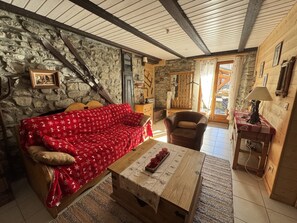 Ein gemütlicher Raum mit einer Steinwand, einem roten, gemusterten Sofa, einem Holztisch mit einer Platte voller roter Tassen, einem braunen Ledersessel und einem Holzschrank mit einer Lampe.