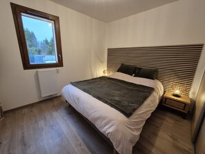 A bedroom with a large bed, a window, a radiator, and a bedside table with a lamp. - New 2-bedroom apartment with parking, ground floor (Arâches-la-Frasse)