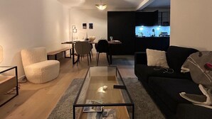 Uma sala de estar moderna com uma mesa de centro de vidro, um sofá preto e uma poltrona branca. Há uma área de jantar com mesa e cadeiras, e uma cozinha visível ao fundo.