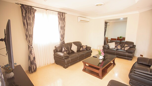 A living room with two leather sofas, a coffee table, a TV, and a dining area in the background. - Luxurious Three Bedroom House at Trassaco East Legon (Madina)