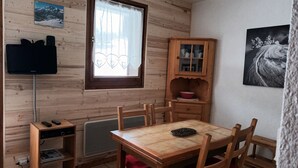 Een kleine, gezellige kamer met een houten wand, een raam met gordijn, een aan de muur bevestigde televisie, een houten kast met glazen deuren en een eettafel met stoelen.