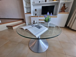 A glass-top table with an open book, a potted plant, and a cup on a tiled floor. - Cozy apartment in L'Escala near the beach (L'Escala)