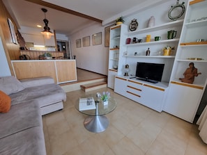 A living room with a sofa, a glass coffee table, a TV on a white entertainment unit, and a kitchen in the background. - Cozy apartment in L'Escala near the beach (L'Escala)