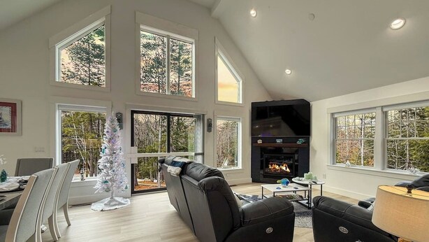 A spacious living room with a fireplace, large windows, and a dining area with a Christmas tree. - Northview Chalet | Brand New, Hot Tub, Spacious (Conway)