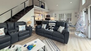 A spacious living room with a black leather sofa, a coffee table, and a staircase leading to an upper level. A white Christmas tree is visible in the background. - Northview Chalet | Brand New, Hot Tub, Spacious (Conway)