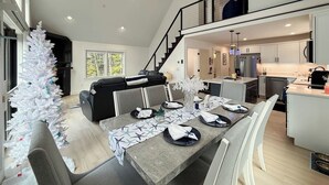 A modern dining area with a high ceiling, a staircase leading to an upper level, and a kitchen in the background. - Northview Chalet | Brand New, Hot Tub, Spacious (Conway)