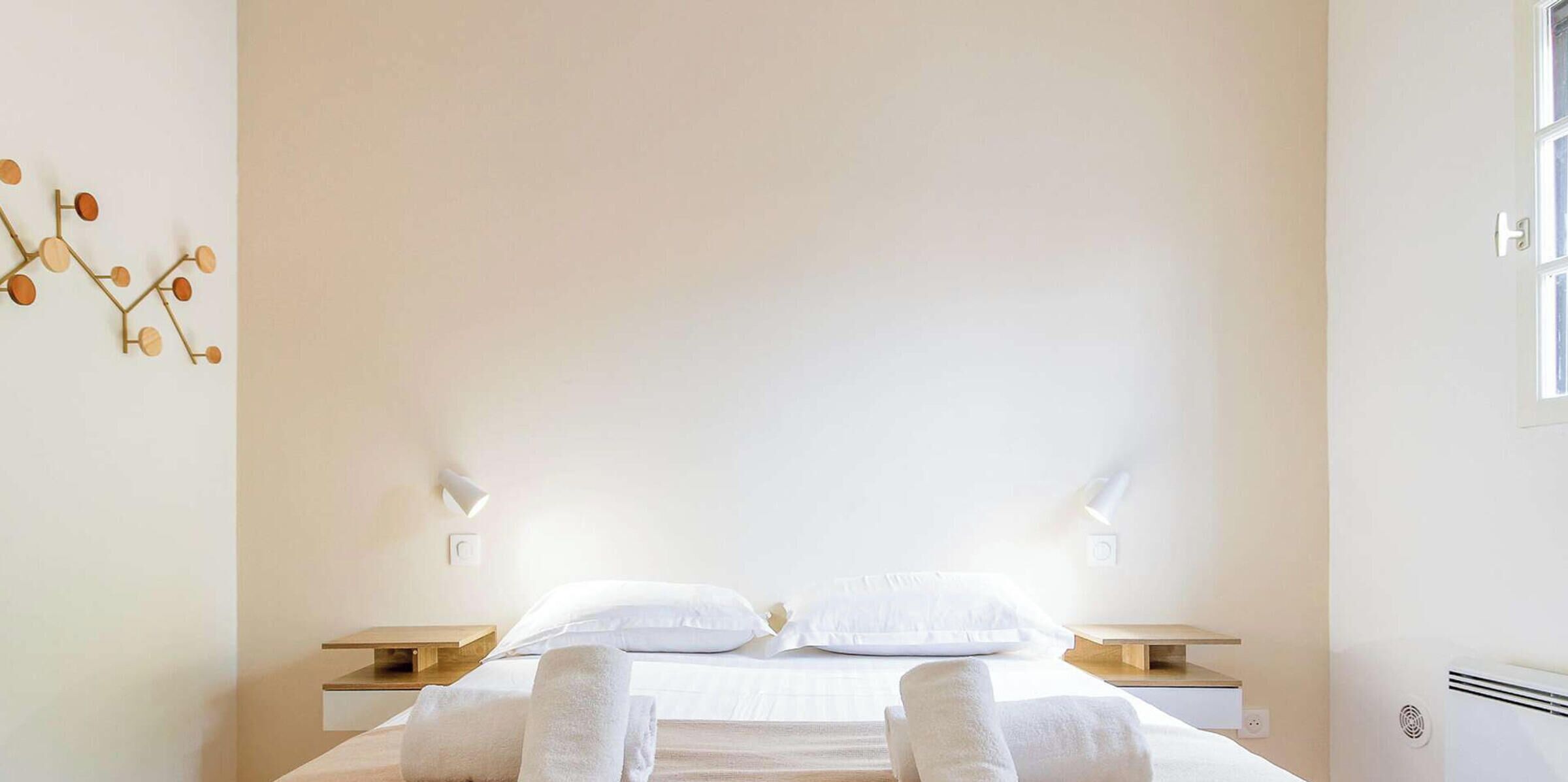 A minimalist hotel room with a large bed, two bedside tables, and a wall-mounted light fixture.