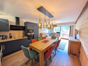 A modern kitchen with built-in appliances, a dining area with a wooden table and colorful chairs, and a view of the outdoors through large windows. - Stunning new apartment 8 pers, 55m² south terrace, near slopes (Montgenèvre)