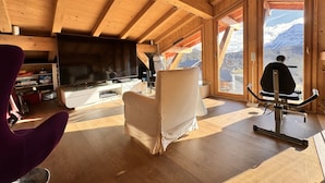 Ein Zimmer mit hohen Decken, Holzboden, einem großen Fernseher, einem Trainingsgerät und Blick auf Berge.