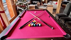 Ein Billardtisch mit rosa Filzbezug, Billardstöcke und Kugeln in einem Raum, der durch einen Weihnachtsbaum und ein Fenster gekennzeichnet ist.