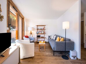 Ein modernes Wohnzimmer mit großem Fenster, einem grauen Sofa, einem weißen Sessel, einem Holztisch und einem Bücherregal.