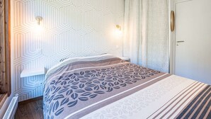 Ein Schlafzimmer mit einer Wand im geometrischen Muster, einem Bett mit Bettdecke im Blattmuster, zwei Wandleuchten und einer Tür mit rundem Spiegel.