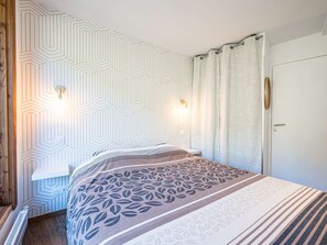 Ein Schlafzimmer mit einer Wand im geometrischen Muster, einem Bett mit Bettdecke im Blattmuster, zwei Wandleuchten und einer Tür mit rundem Spiegel.
