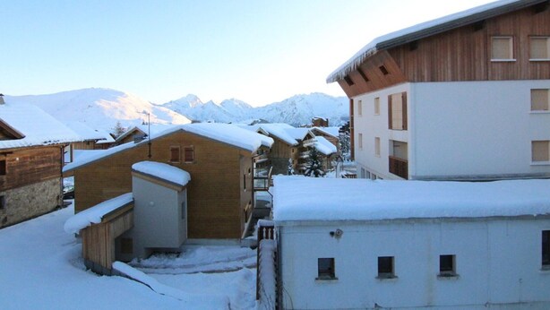 Ein schneebedecktes Gebäude mit Holzfassade und weißem Oberteil, vor einer Kulisse schneebedeckter Berge.