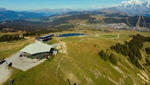 A modern building with a flat roof and a large parking area, surrounded by a mountainous landscape and a small town in the distance.