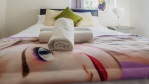 A neatly made bed with a rolled towel, pillows, and a blanket with a face design.