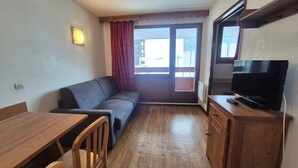 Ein kleiner, möblierter Raum mit einem Holztisch, einer Couch, einem Fernseher und einem Fenster mit Vorhängen.