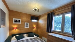 A cozy room with wooden walls, a bed with a fluffy blanket, two wall-mounted lamps, and a window with curtains. - 3-Room Alcove Apartment for 6 in Méribel, near shops (Méribel)