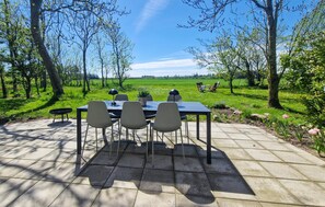 Uma área de refeições ao ar livre com mesa e cadeiras, cercada por árvores e um campo gramado.