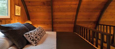 Habitación de cabaña de madera con cama, almohadas, una ventana y barandilla de madera.