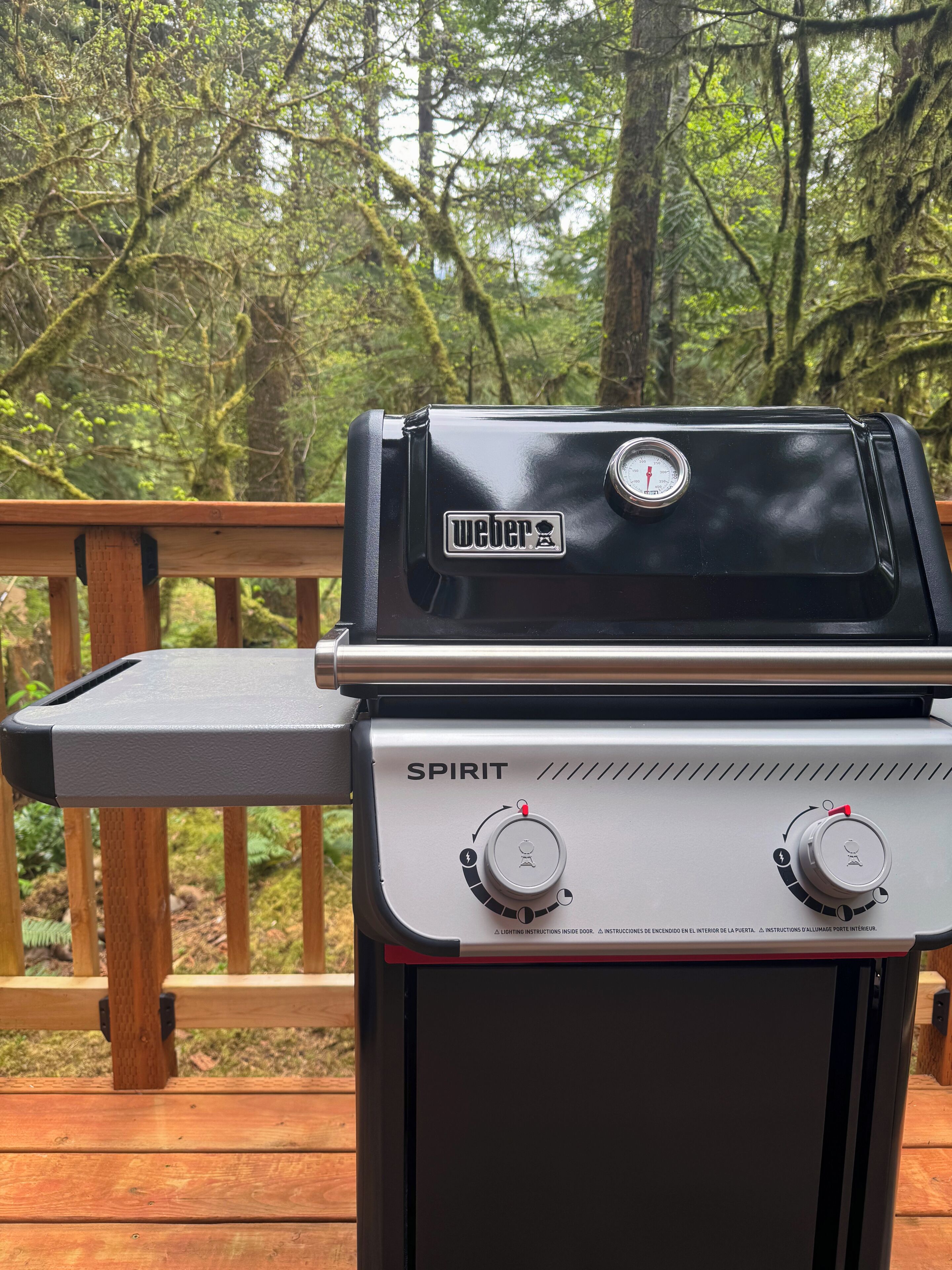 A Weber Spirit grill with a digital thermometer, set on a wooden deck with a forest backdrop.