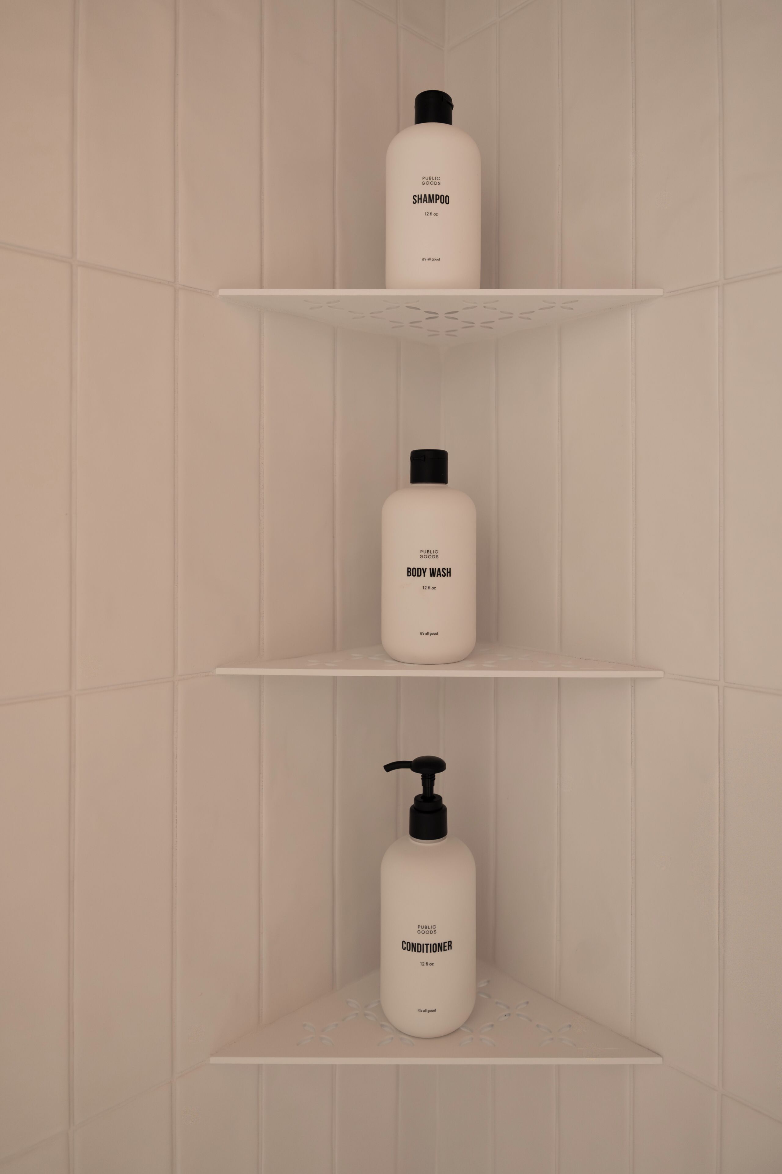 A bathroom with three white shelves holding bottles of Public Goods shampoo, body wash, and conditioner.