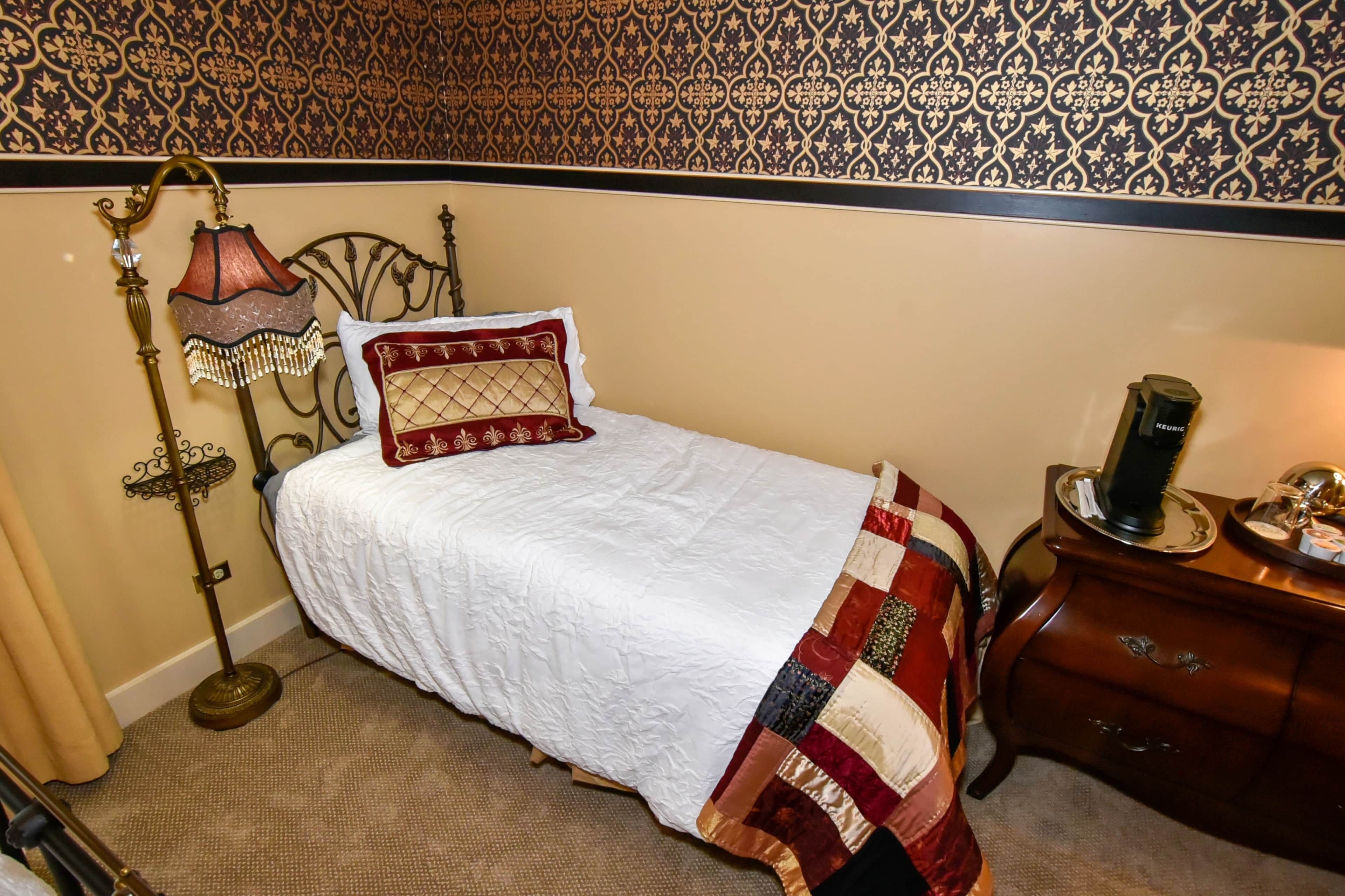 A neatly made bed with a white comforter and a red and gold quilt, a vintage-style lamp, and a wooden dresser with a mirror and a small table.