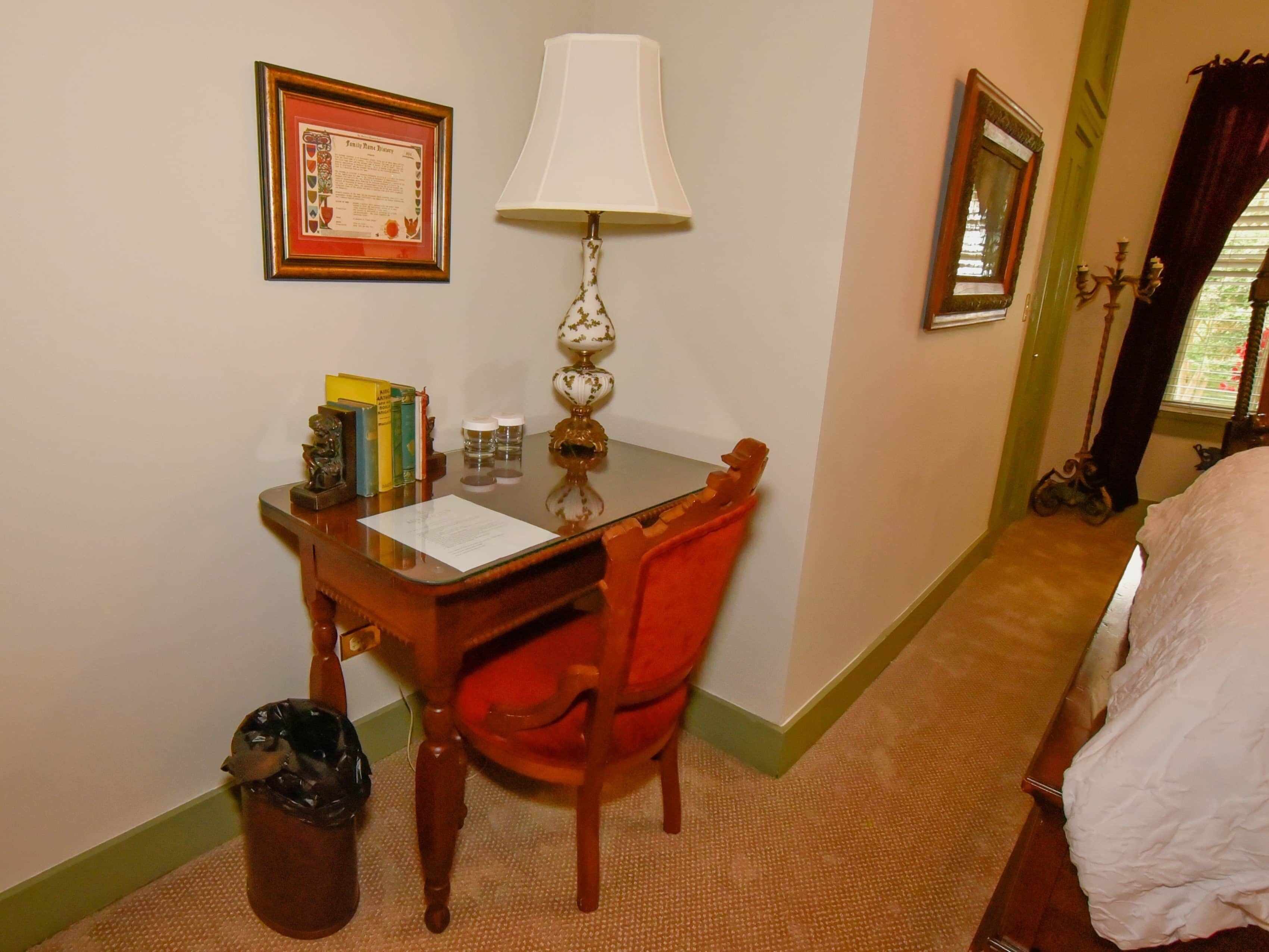 A hotel room with a wooden desk, a red chair, a lamp, a framed certificate on the wall, and a bed with a white cover.