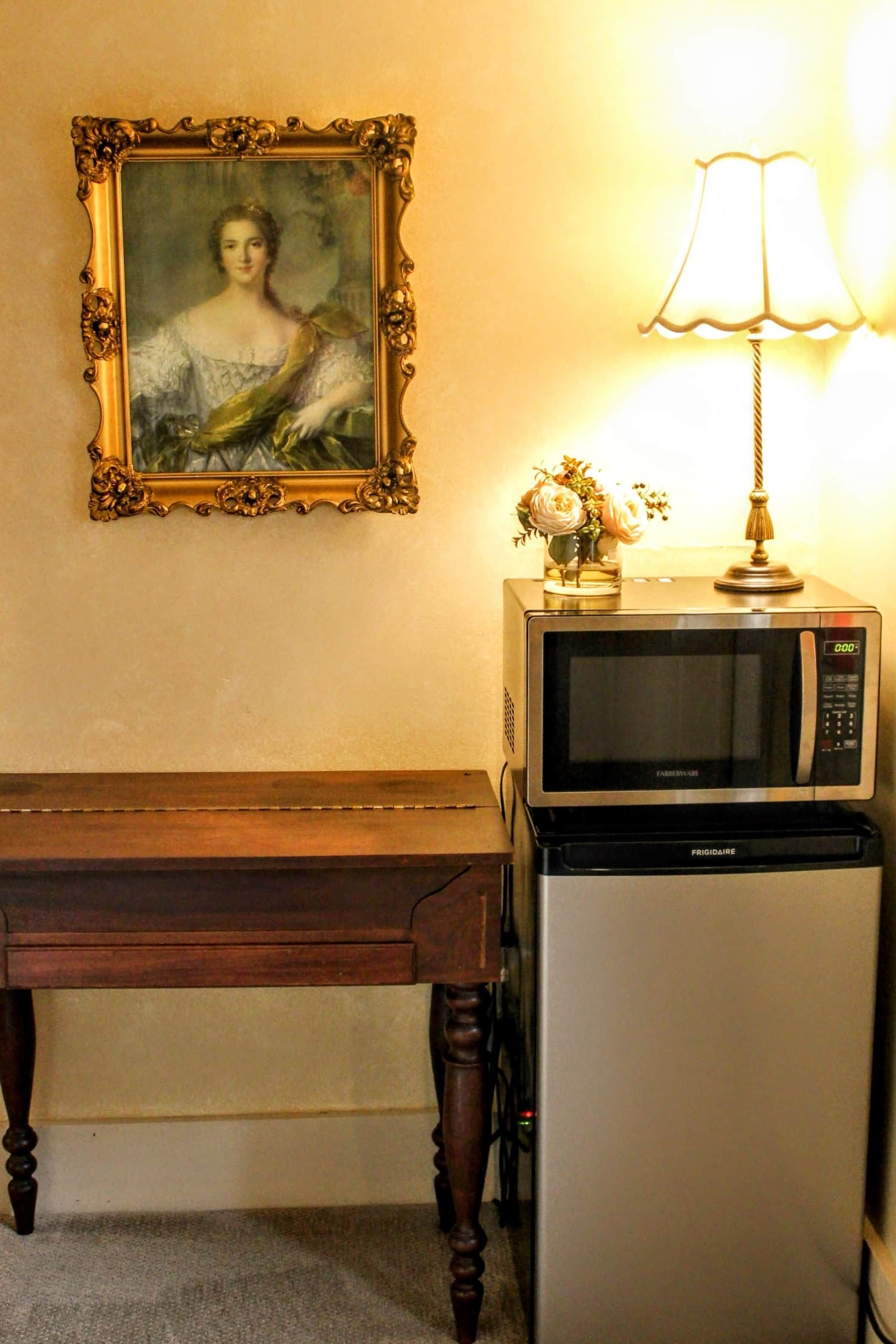 Una habitación con una mesa de madera, un retrato en un marco en la pared, una lámpara y un microondas.
