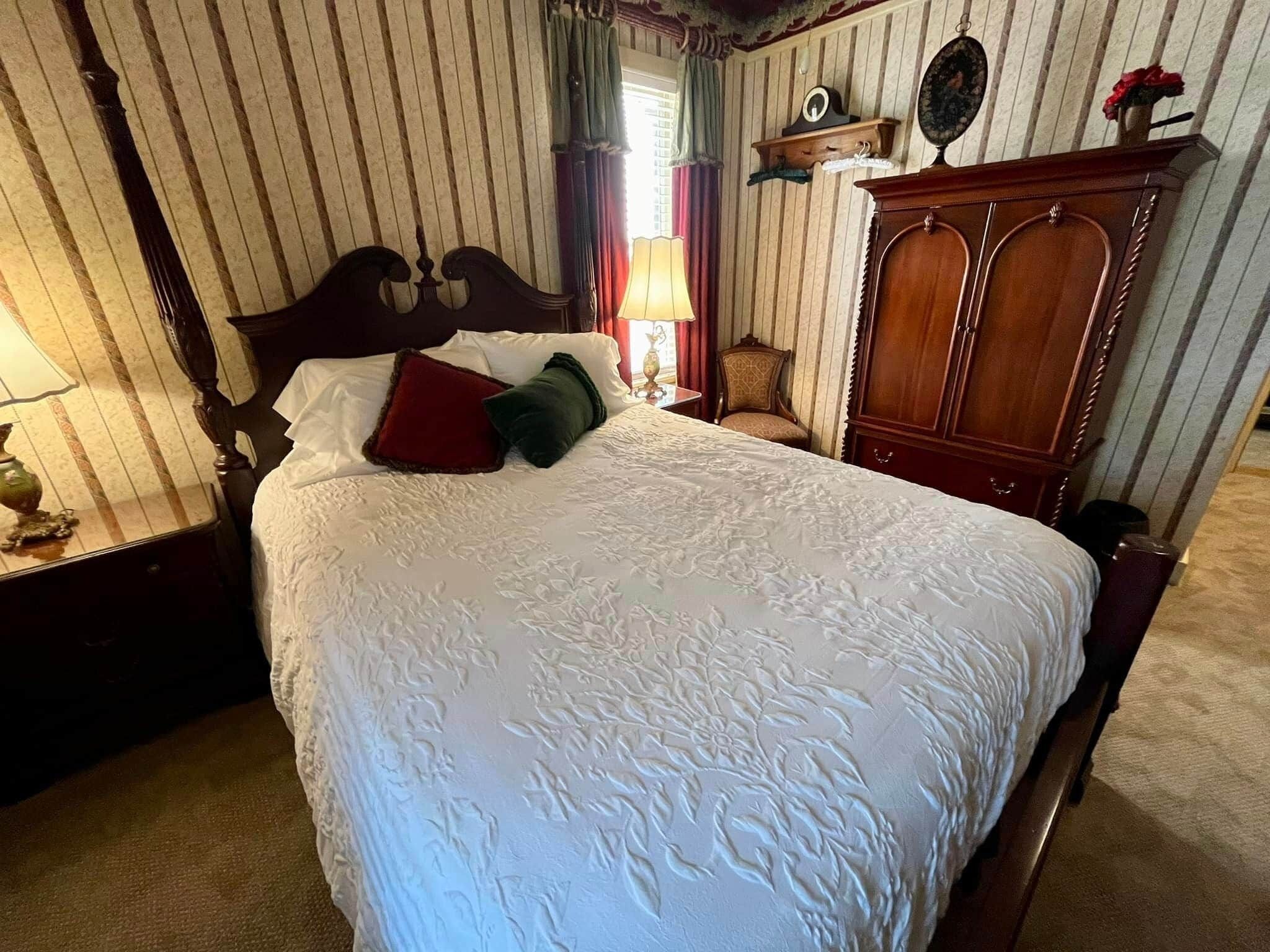 A neatly made bed with a floral-patterned bedspread, a wooden nightstand with a lamp, a wooden wardrobe, and a chair in a room with striped wallpaper.