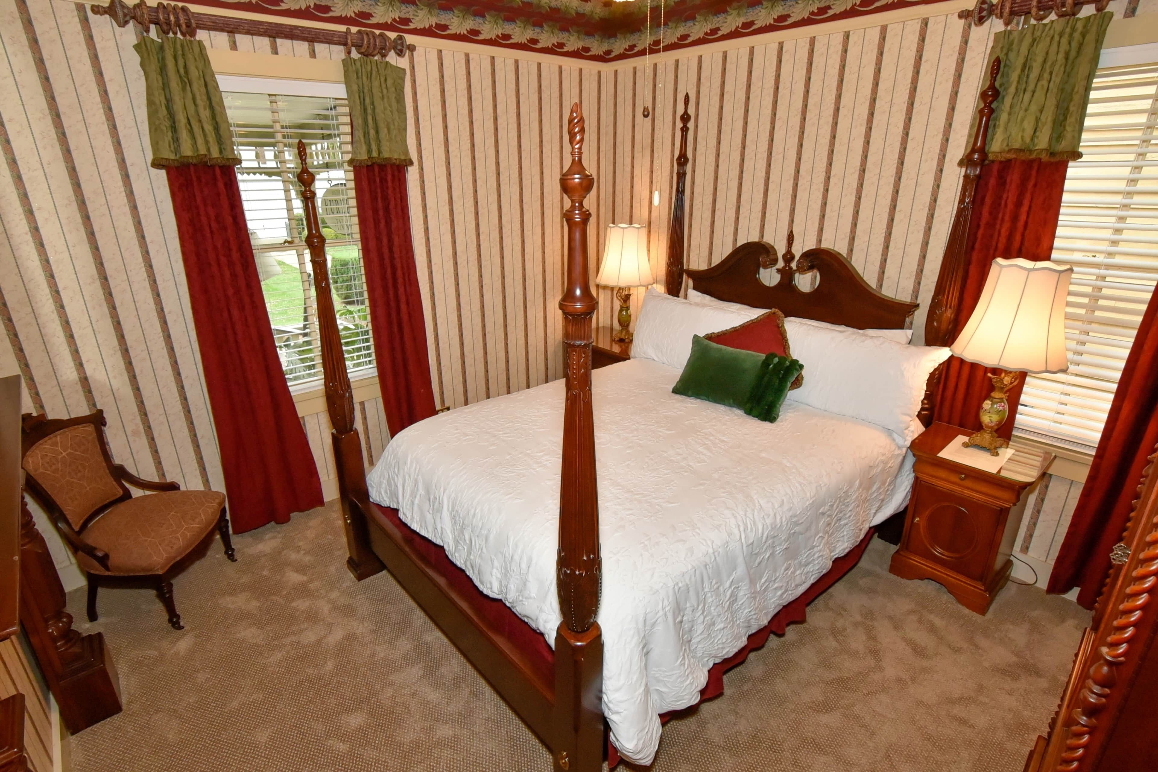A four-poster bed with a wooden headboard, two bedside tables with lamps, a chair, and striped wallpaper.