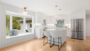Eine moderne Küche mit weißen Schränken, einer großen Kochinsel mit Barhocker, einem Edelstahl-Kühlschrank und Blick durch große Fenster.
