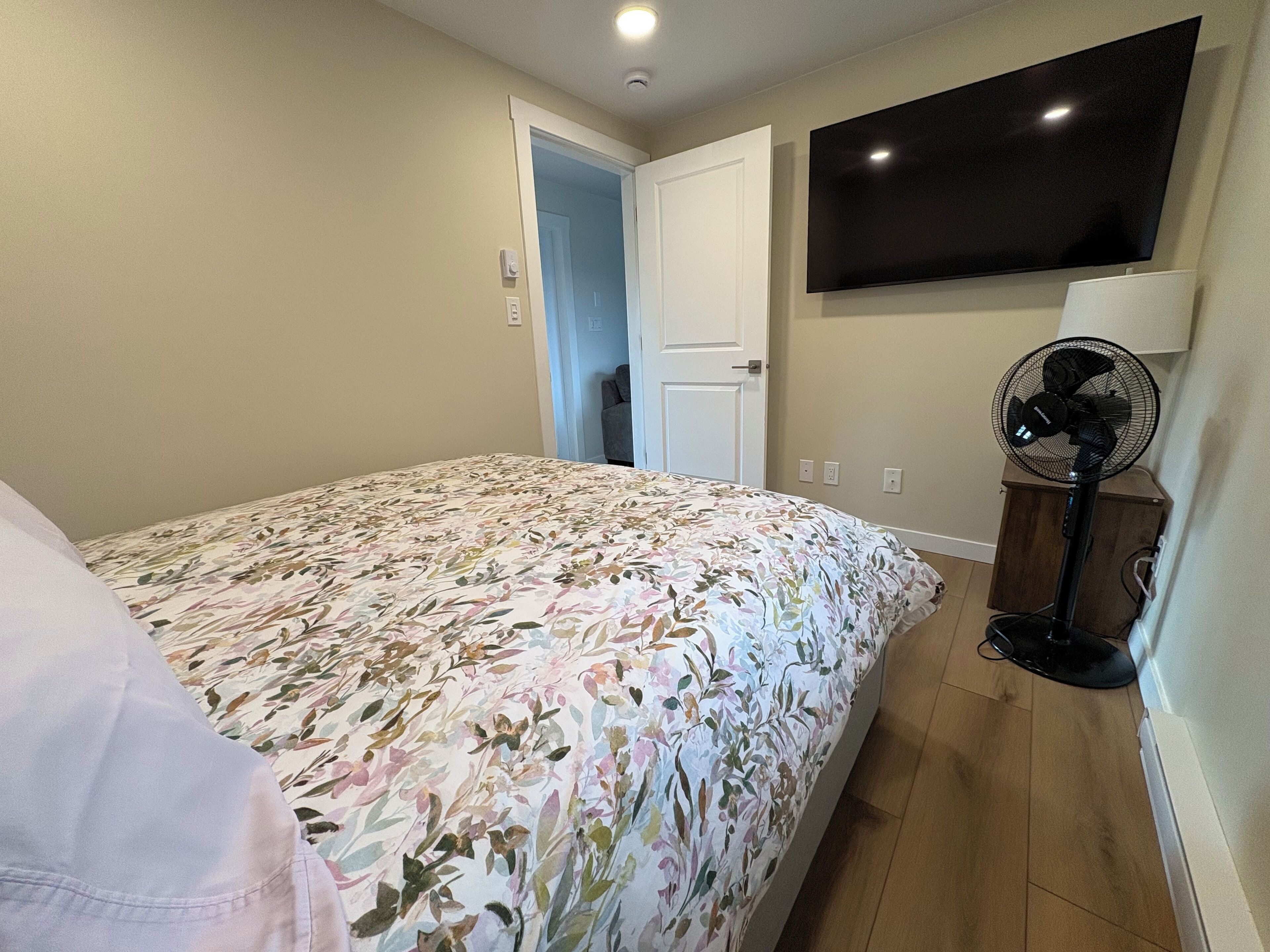 A bedroom with a floral bedspread, a TV mounted on the wall, and a standing fan.