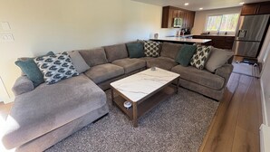 A modern living room with a sectional sofa, a marble coffee table, and a kitchen area with wooden cabinets and stainless steel appliances.