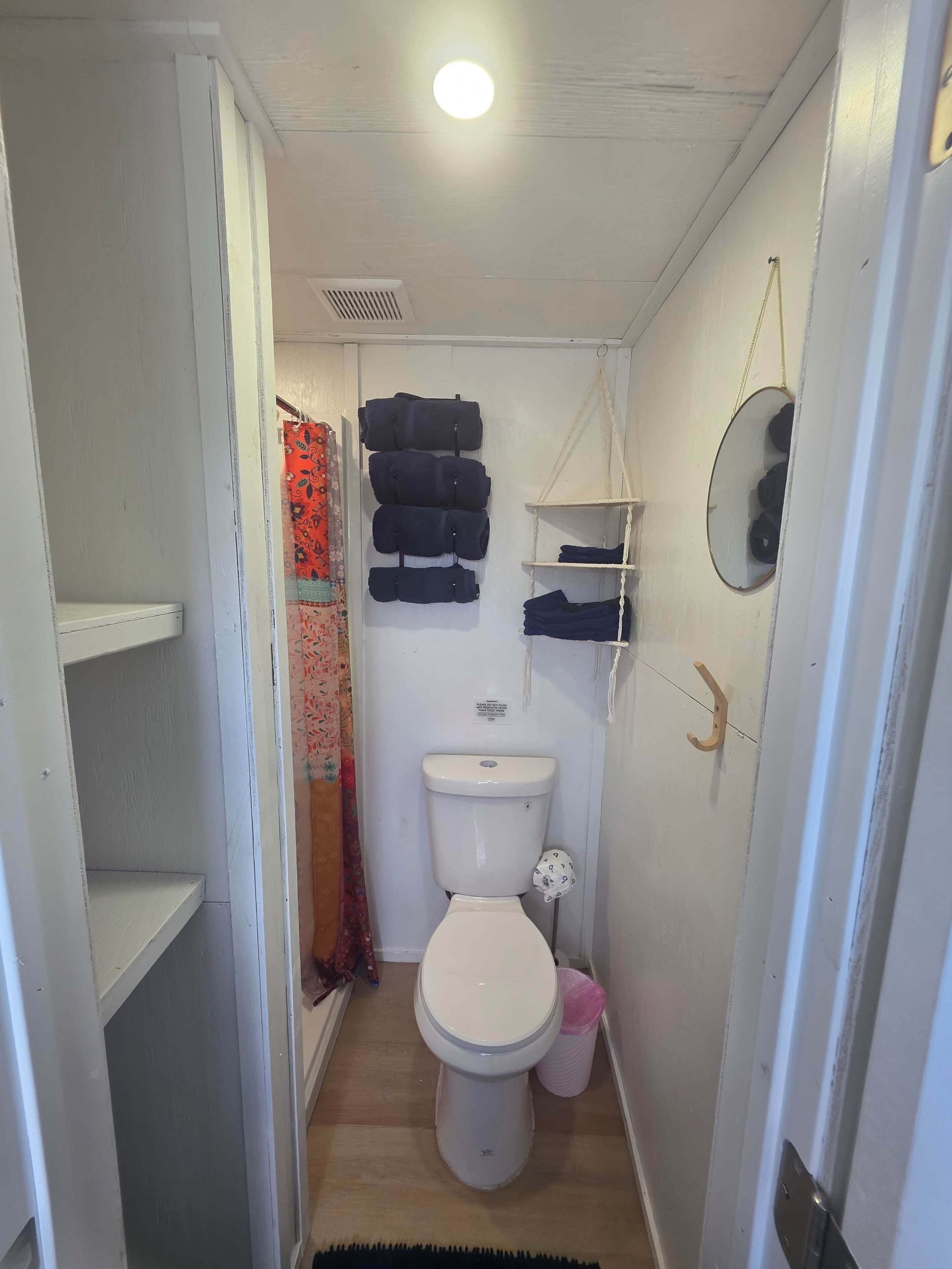 A small, narrow bathroom with a toilet, a hanging toilet paper holder, a shelf with folded clothes, and a mirror on the wall.