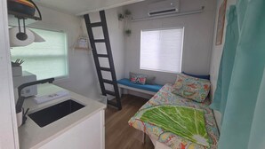 Habitación compacta con kitchenette, cama con colcha colorida, ventana con persianas y unidad de aire acondicionado.