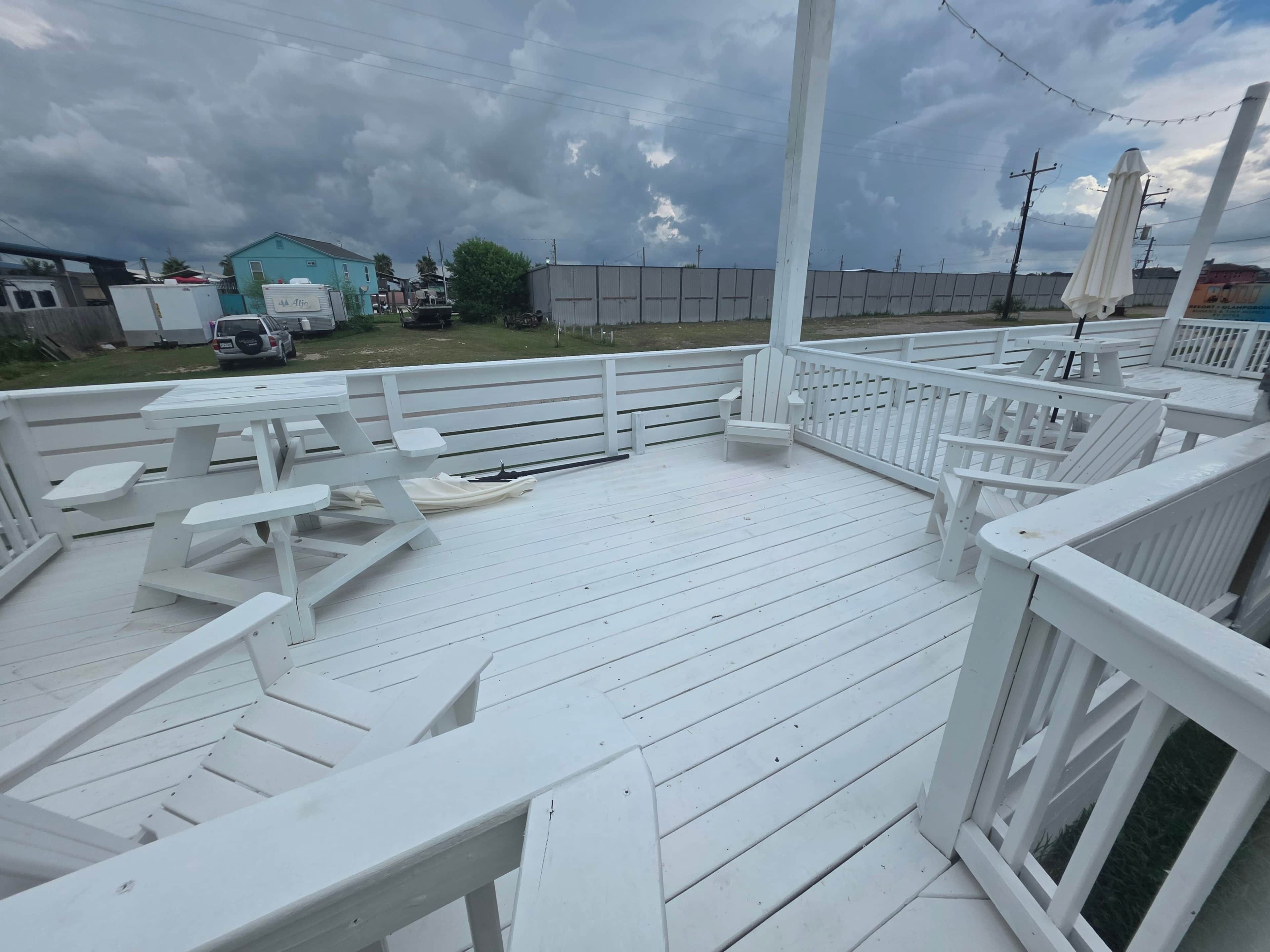 Una terraza techada al aire libre con bancos y mesas blancos, rodeada de barandales blancos.