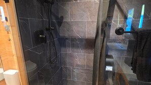 Un bany modern amb dutxa de vidre, rajoles de marbre fosc, un vàter i detalls de fusta.