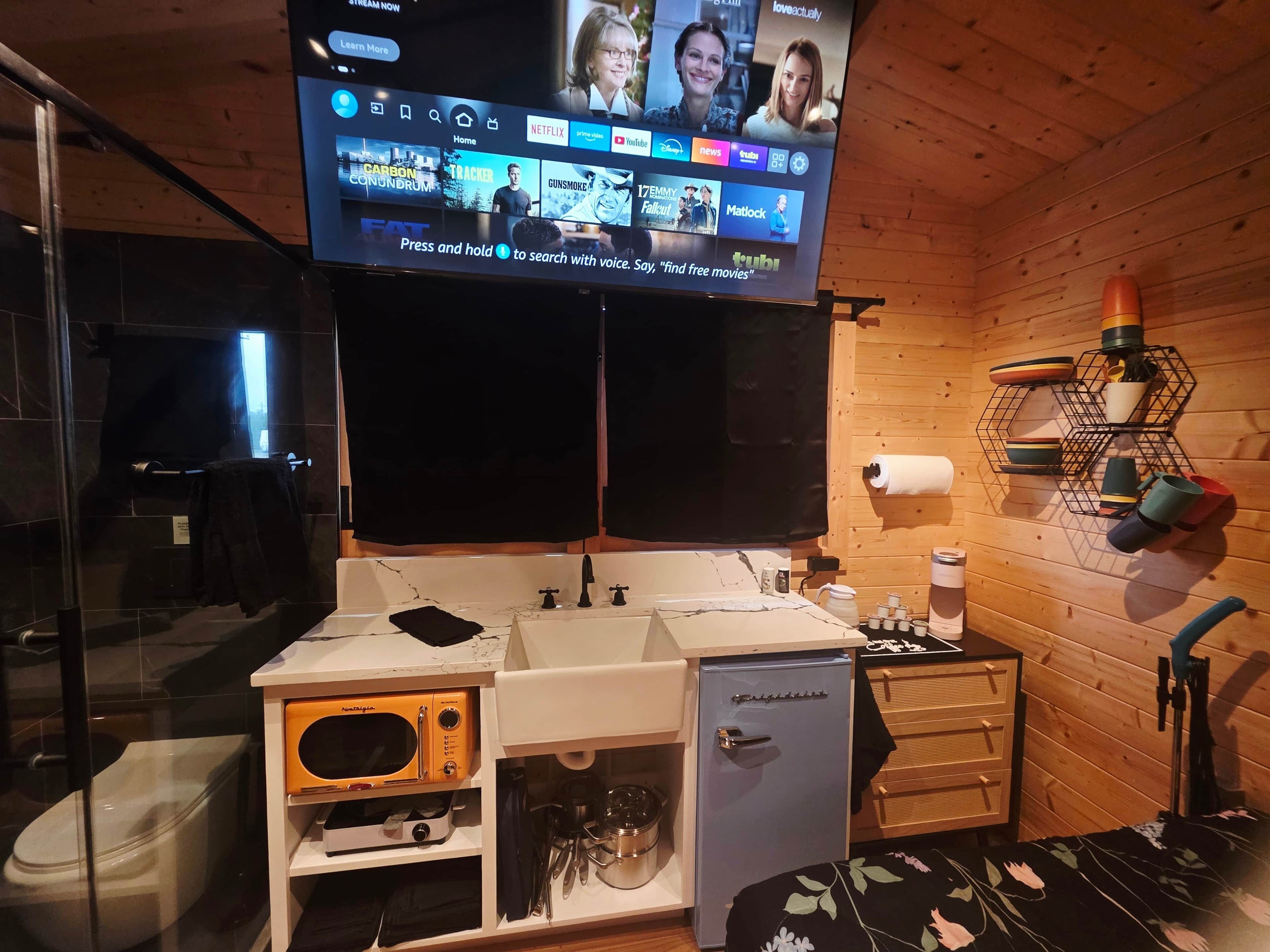 A compact hotel room with a wooden interior, a television mounted on the wall, a sink with a mirror, a small refrigerator, and a microwave.