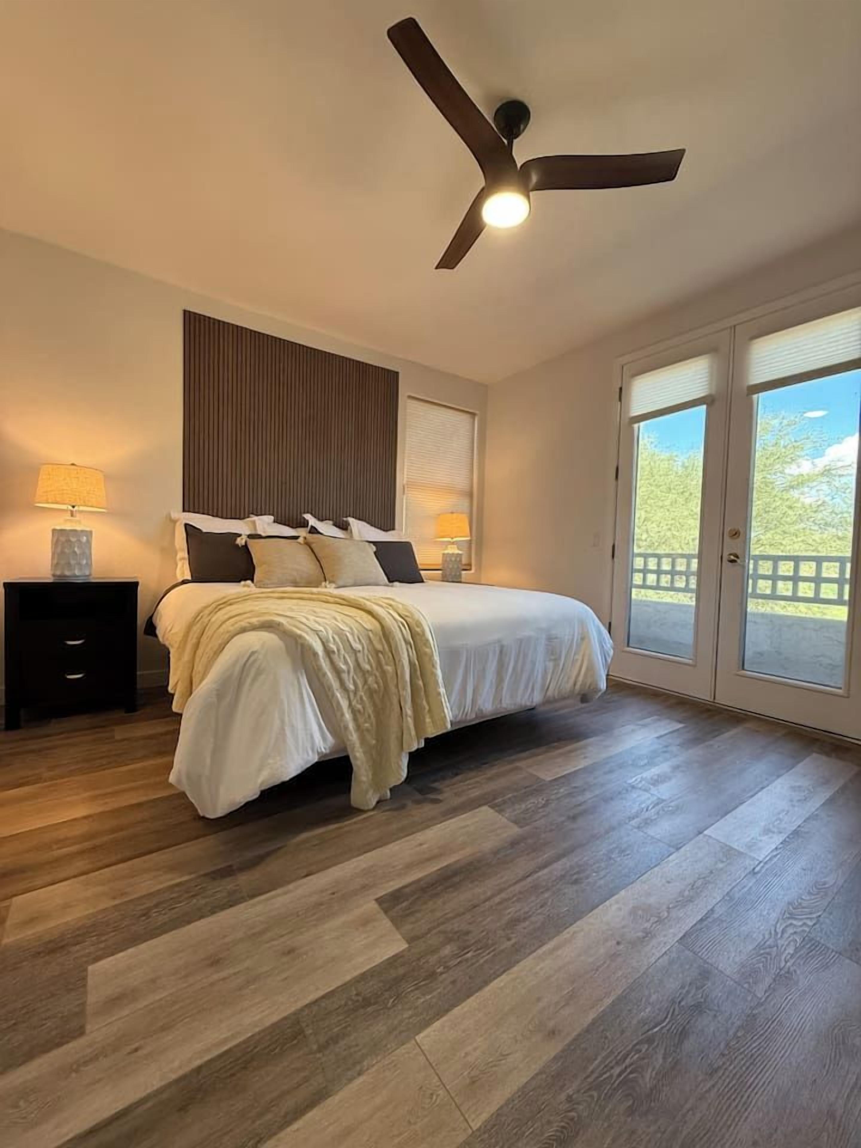 A bedroom with a bed, a ceiling fan, a nightstand, and a window with a view.