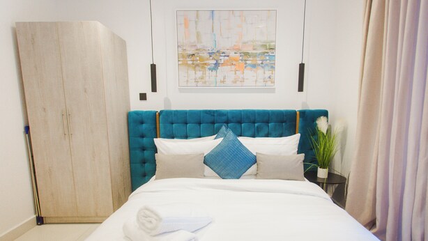 A neatly made bed with white linens and a folded towel, a teal upholstered headboard, and a painting above the bed. - Serene Studio Apartment at Oasis Park (Accra)