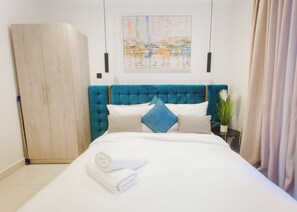 A neatly made bed with white linens and a folded towel, a teal upholstered headboard, and a painting above the bed. - Serene Studio Apartment at Oasis Park (Accra)
