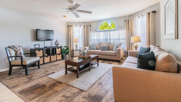 A living room with a sofa, armchair, coffee table, TV, and a ceiling fan. - It's All NEW & Near Disney! Designer Kitchen, Sleeps 6, NO Steps, Patio, Pool (Kissimmee)